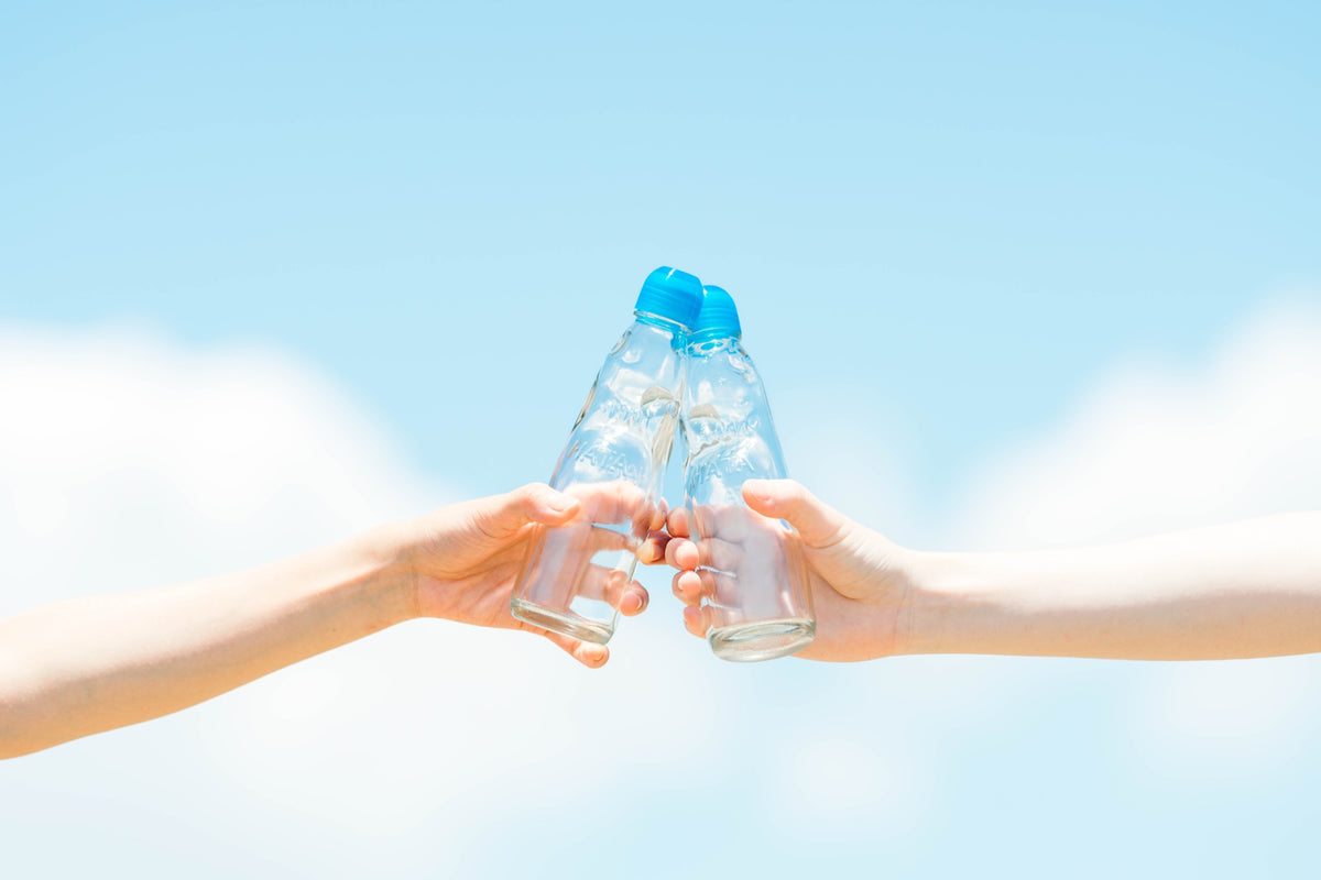 Two hands toasting Ramune-style soda bottles against a blue sky